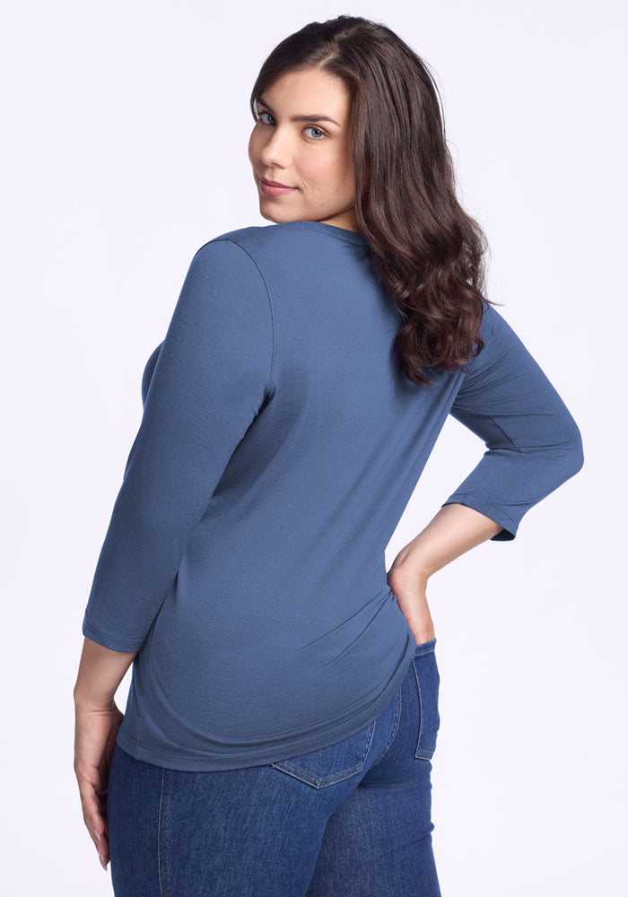 A woman with long brown hair wears the Woolx Jenny 3/4 Sleeve Crew Neck in Skyline and blue jeans, standing turned to the side and looking over her shoulder at the camera against a plain light background.