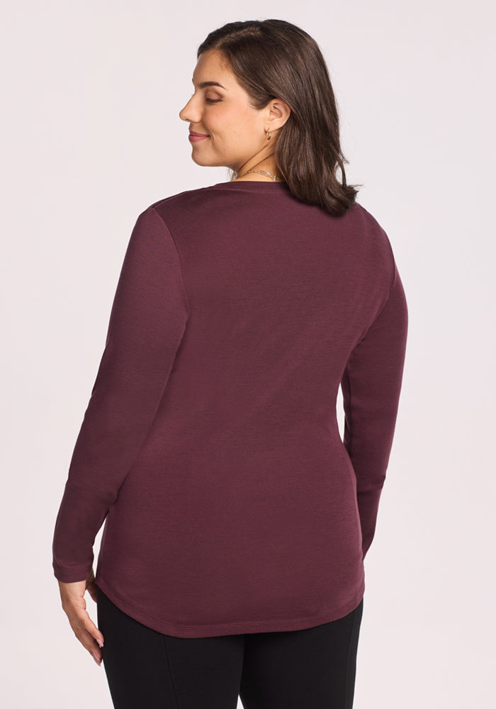 A woman with short brown hair stands with her back to the camera, softly smiling with eyes closed, wearing the Woolx Eva Tunic in Chocolate Fig and black pants against a plain light background.