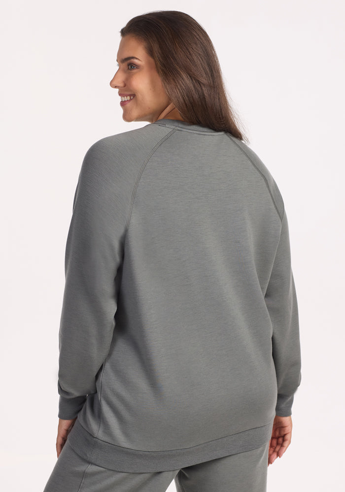 A woman with long brown hair smiles while looking to the side, wearing the Woolx Bailey Crewneck Sweatshirt in Sedona Sage with matching pants. She is shown from the back against a plain light background.