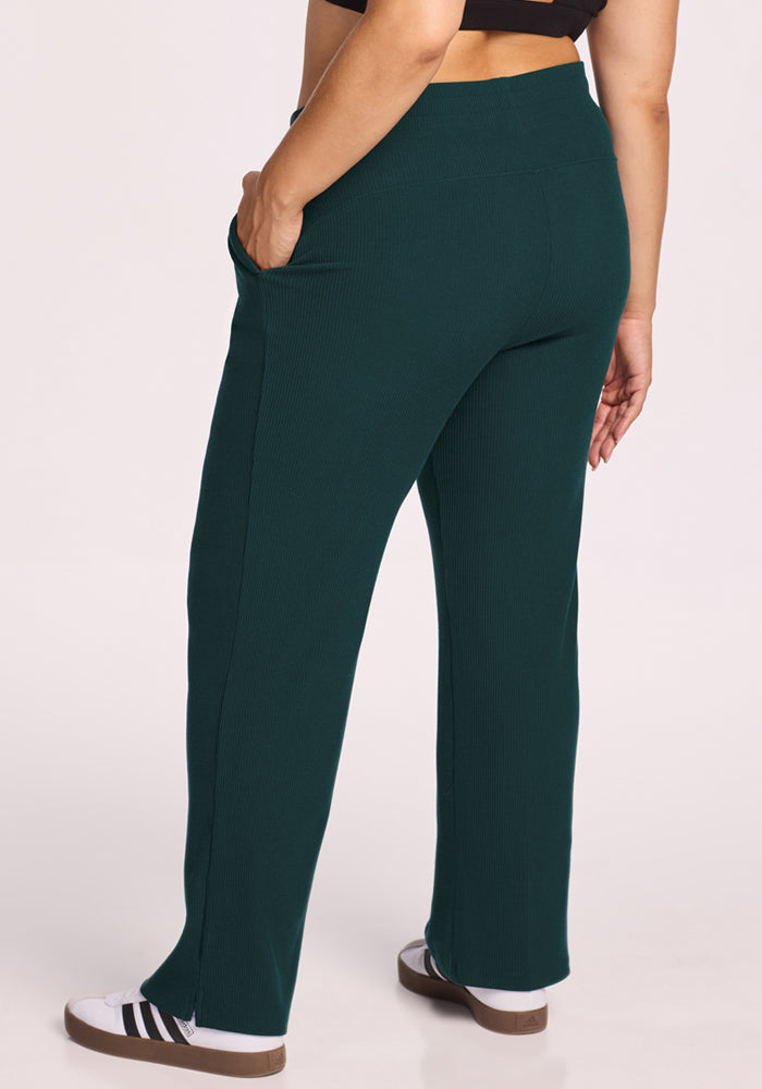 Someone stands against a plain background with one hand in their pocket, wearing Woolx's Luca Ribbed Pants in Deep Timber, styled with a black crop top and white sneakers featuring black stripes.