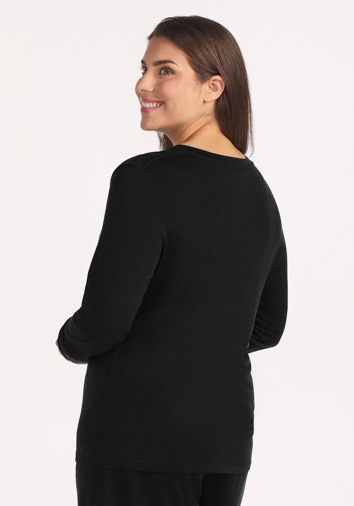 A woman with long brown hair smiles over her shoulder, wearing the Woolx Lily V Neck Lounge Top in Black. She stands against a white background, ready for restful nights or cozy travels.