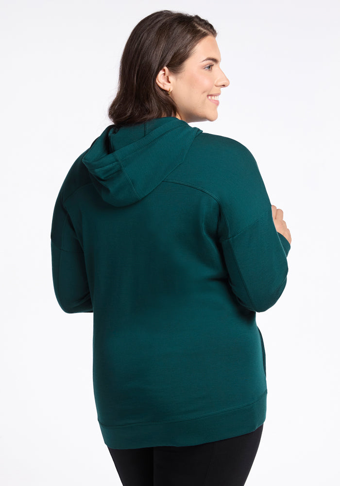 A woman with shoulder-length brown hair, facing away, wears the Woolx Callie Hoodie in Riverbank Green and black pants, against a plain white background.