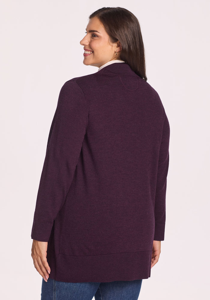 A woman with long brown hair, wearing the Woolx Macie Sweater in Deep Plum over a white top and blue jeans, smiles and looks over her shoulder against a plain light background.