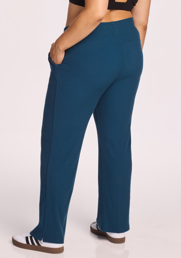 Someone wearing Woolx's Luca Ribbed Pants in Majolica Blue, a black crop top, and black-and-white sneakers stands with one hand in their pocket against a plain light background. The ribbed pants add texture to the relaxed look.