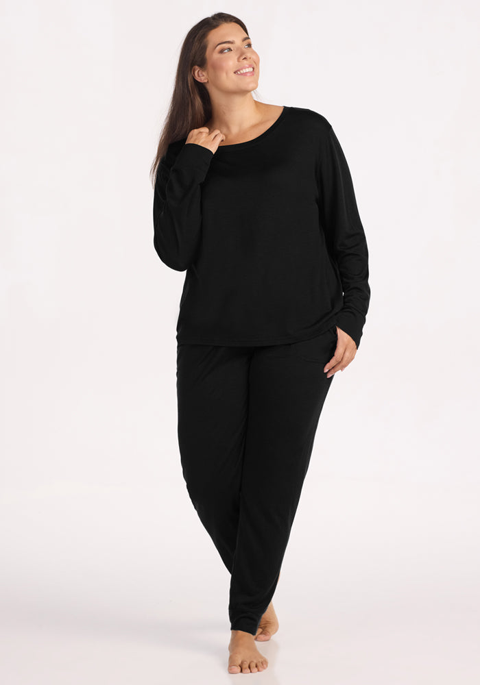 A woman in Woolx Mary Kate Lounge Pants in black stands barefoot on a white background, smiling and looking to the side—ideal for stylish and comfortable travel lounge wear.