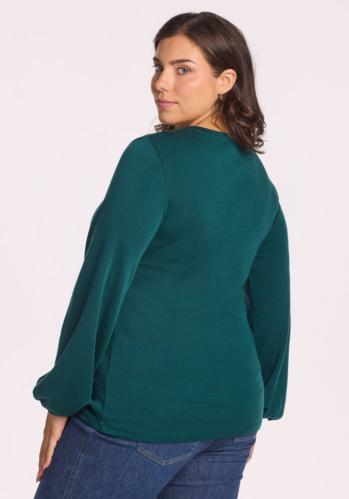 A woman with dark hair, dressed in the Woolx Charlotte Puff Sleeve Top in Riverbank Green and blue jeans, stands looking over her shoulder at the camera against a plain light background.