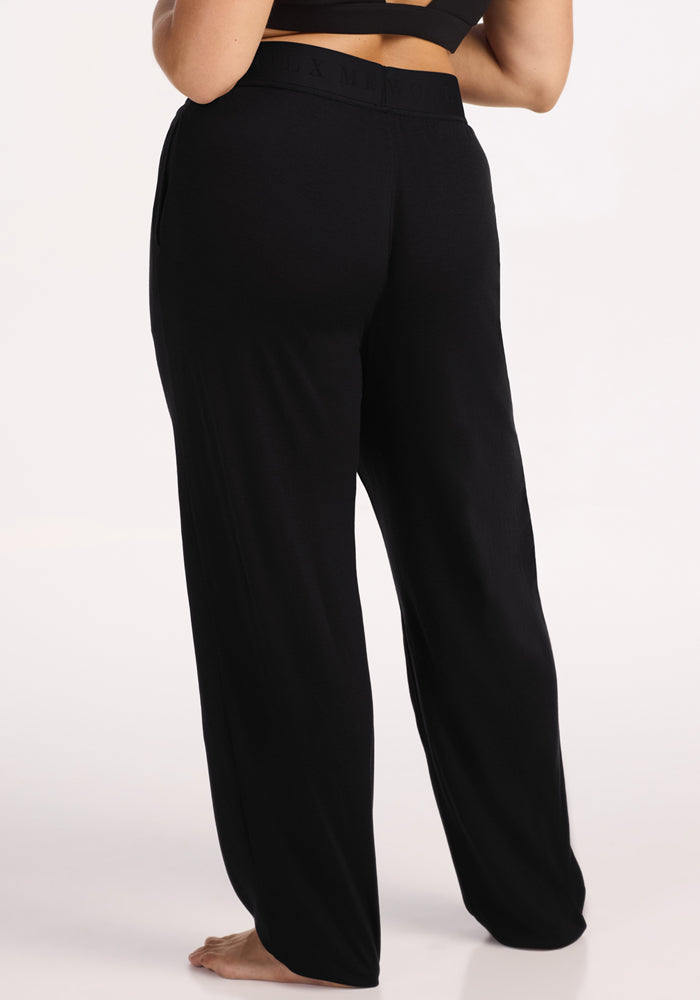 A person stands barefoot, viewed from behind, wearing Woolx Maya Lounge Pants - Black and a black crop top against a plain light background.