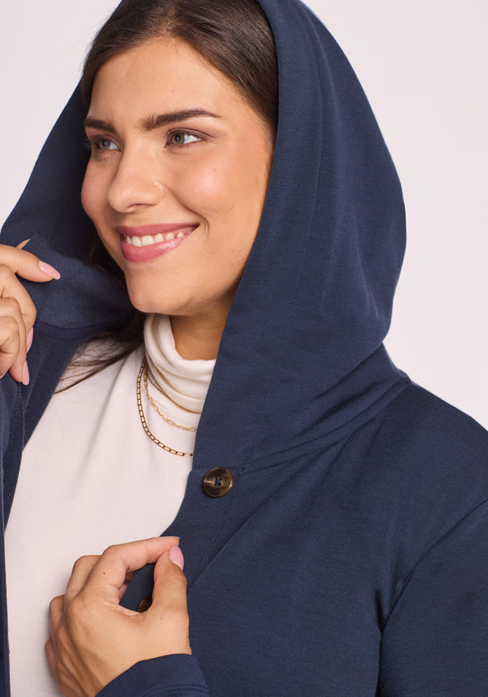 A woman smiles as she lifts the hood of her Woolx Edna Hooded Cardigan in Deep Navy, layering it over a white turtleneck and accessorizing with a gold chain necklace.