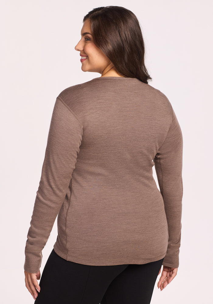 A woman with long brown hair smiles while facing away from the camera, wearing the Woolx Hannah Long Sleeve Top in Simply Taupe and black pants against a plain light background.