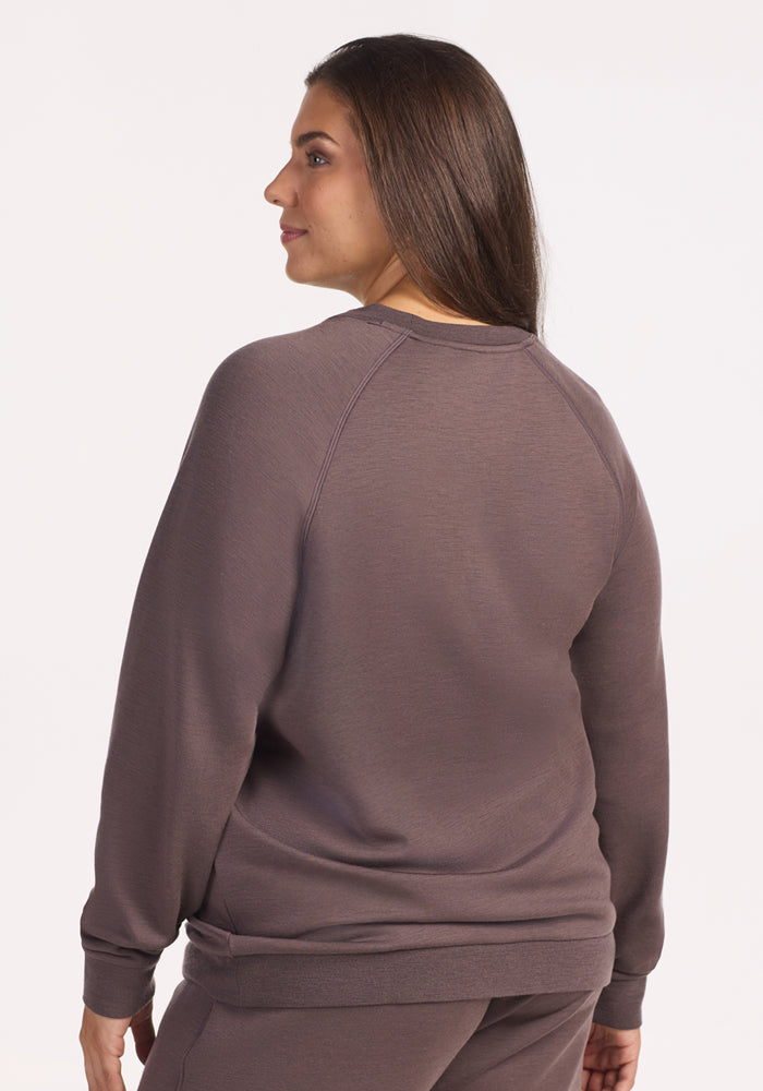 A woman with long brown hair wears the Woolx Bailey Crewneck Sweatshirt in Plum Truffle and matching pants, standing against a plain light background and looking over her shoulder.