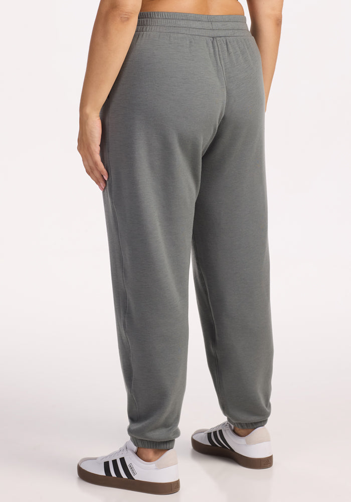 A person is standing facing away, wearing Woolx Parker Sweatpants in Sedona Sage and white sneakers with black stripes, against a plain white background.