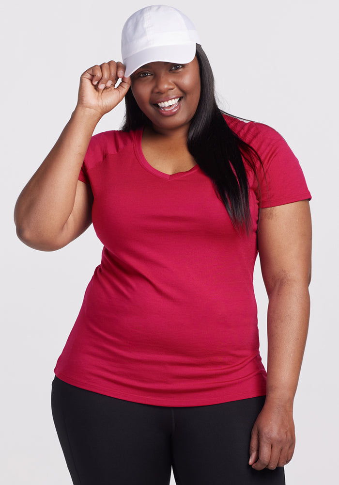 A smiling woman in a Woolx Mia V Neck - Poppy Spark shirt, white baseball cap, and black pants stands against a light background, holding her cap's brim with one hand. 