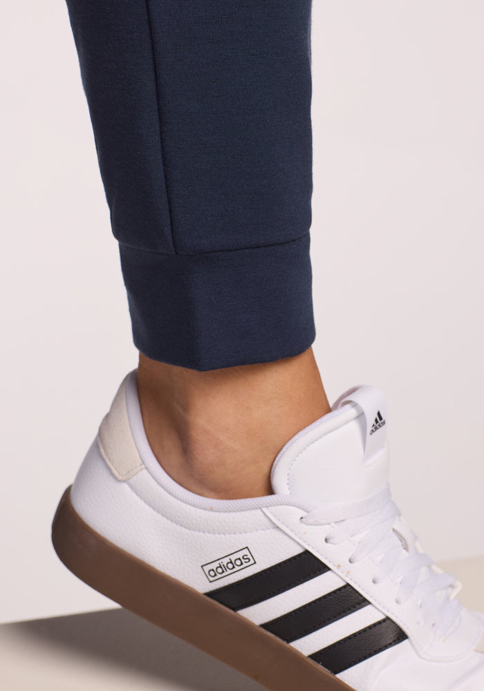 Close-up of a person wearing Woolx Willa Joggers in Deep Navy with white Adidas sneakers featuring black stripes and a brown sole, standing on a light-colored surface.