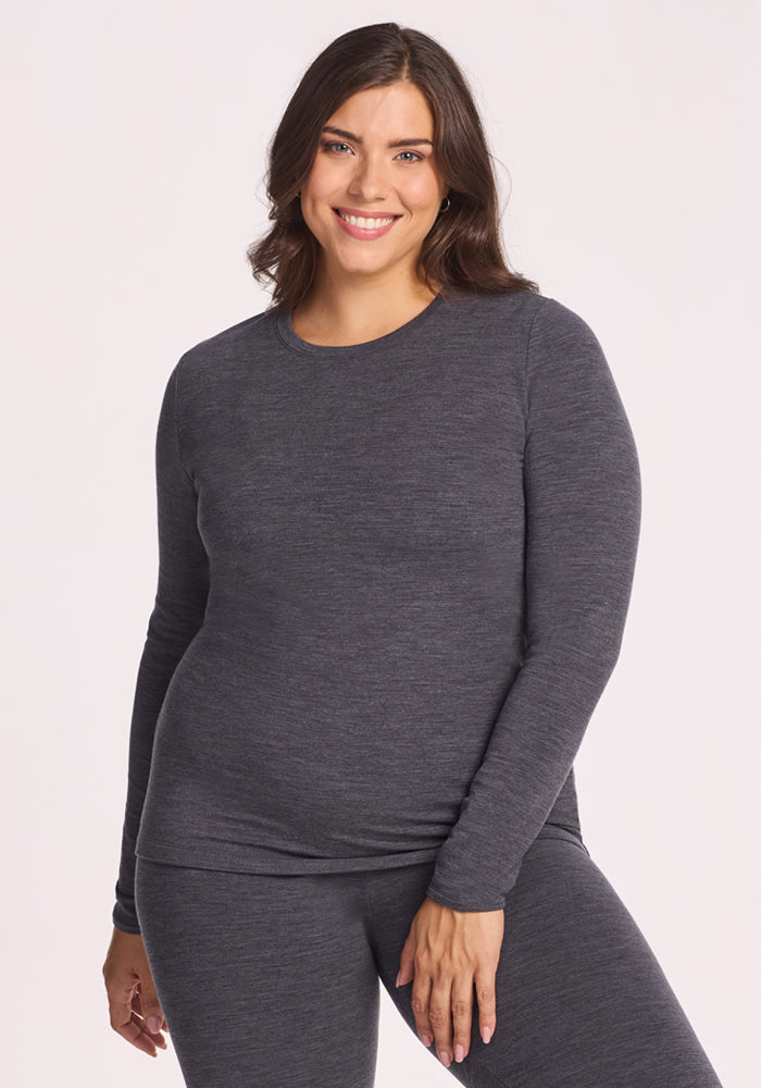 A woman with wavy brown hair smiles while wearing the Woolx Sadie Baselayer Top in Cinder Melange, paired with matching bottoms, standing against a plain light background. 