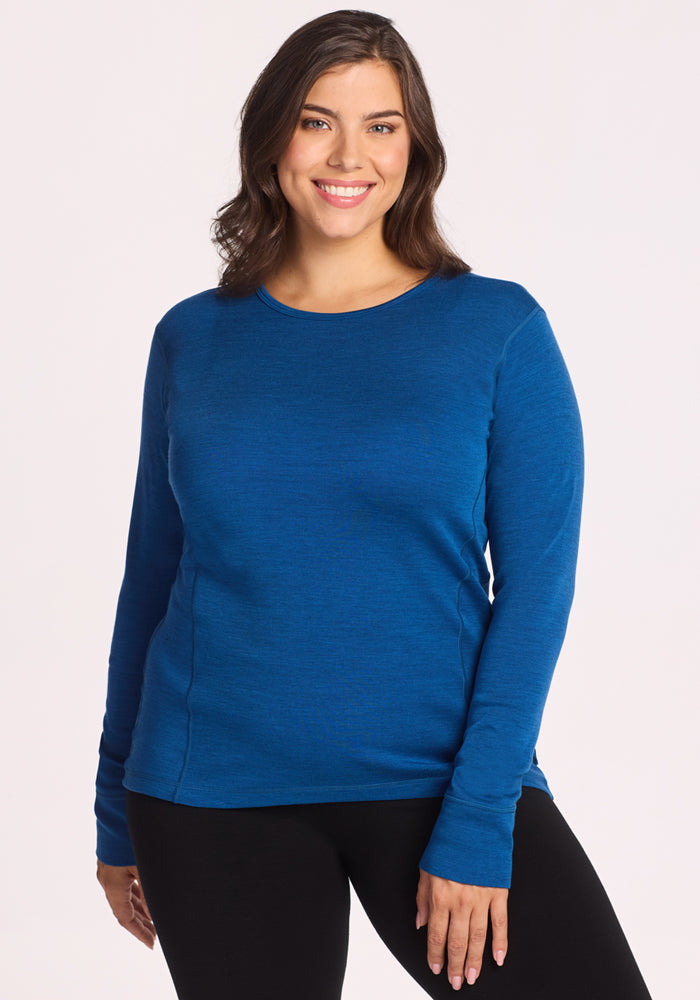 A woman with long brown hair is smiling and wearing the Woolx Hannah Long Sleeve Top in Nightfall, paired with black pants, standing against a plain light background. 