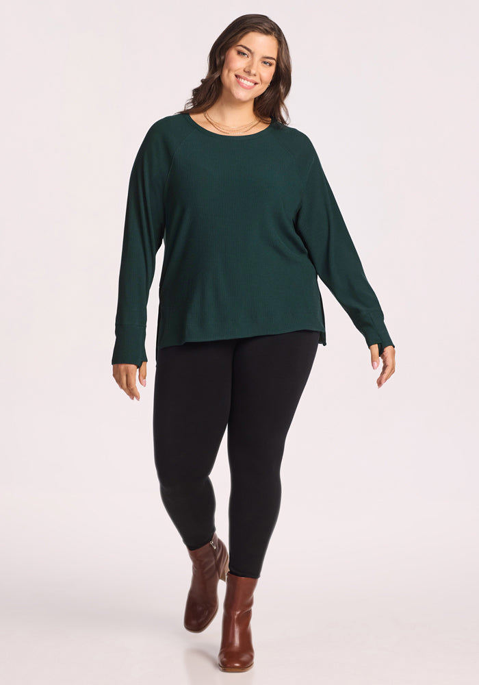 A woman with long brown hair smiles while walking, wearing the Woolx Ainsley Sweater in Deep Timber, black leggings, and brown ankle boots against a light background.