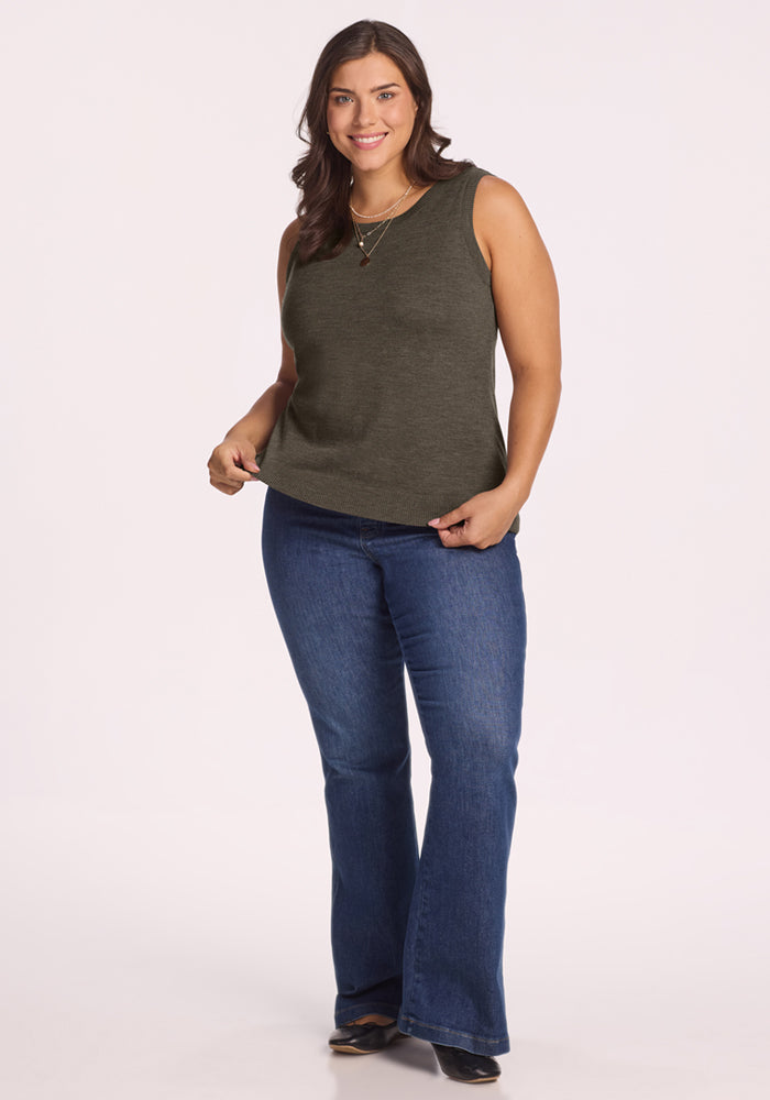 A woman with long brown hair wears the Woolx Chessy Tank Top in Pine Needle, blue bootcut jeans, and black shoes. She stands smiling against a plain light background, holding the hem of her tank top with one hand.