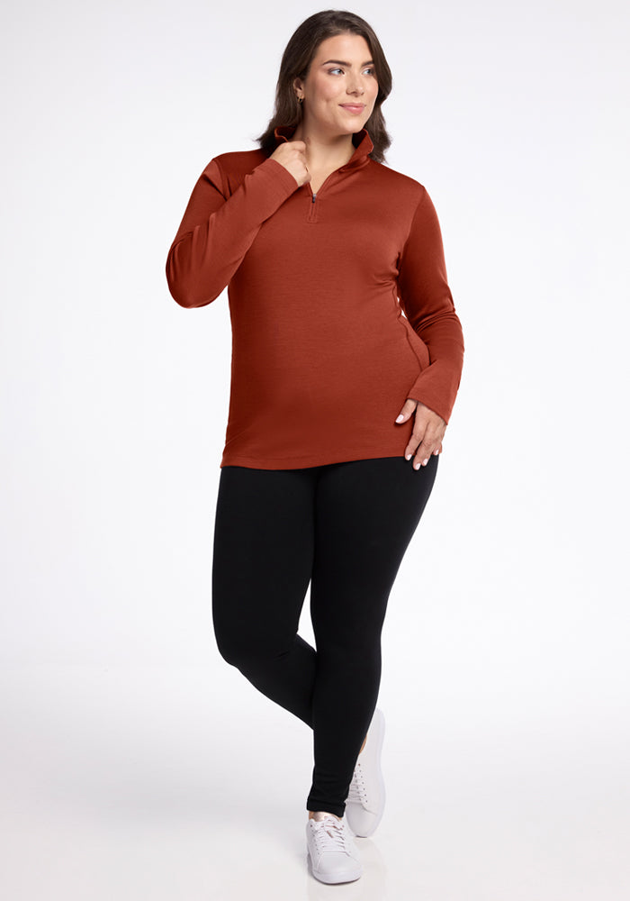 A woman poses in the Woolx Brooke 1/4 Zip - Maple Leaf, a rust-colored long-sleeve top, paired with black leggings and white sneakers. One hand touches her collar while the other rests on her hip against a plain white background.