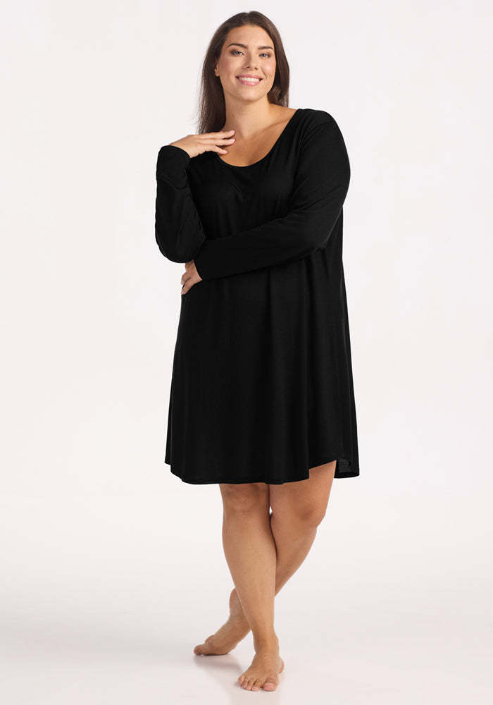 A woman with long brown hair smiles barefoot on a white background, wearing the Woolx Lucille Long Sleeve Nightgown in black and resting one hand on her chin. 