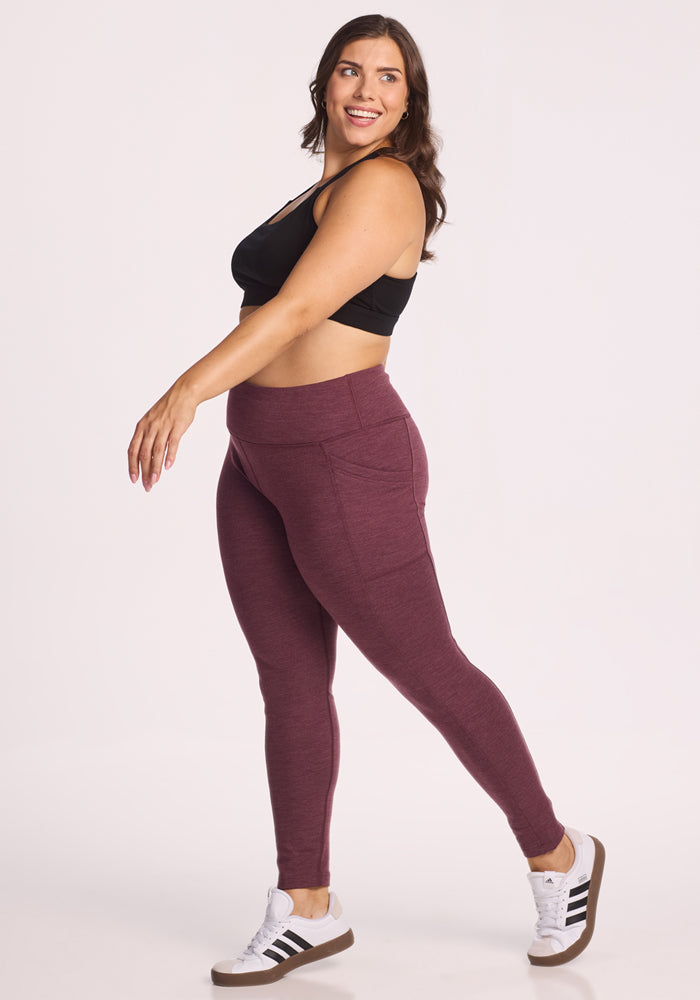 A woman in a black sports bra, Woolx Frost Duralite® Leggings - Grape Harvest Melange, and white sneakers with black stripes smiles while posing with one leg slightly raised against a plain white background.