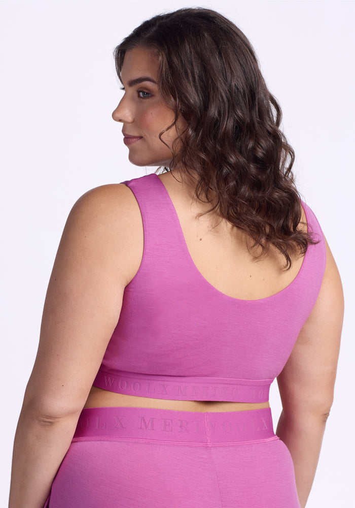 A woman with wavy brown hair wears the Woolx Aubrey Lounge Bra in Wild Blossom, featuring a logo band, and high-waisted leggings, standing slightly turned to display the back and side of her outfit against a plain white background.