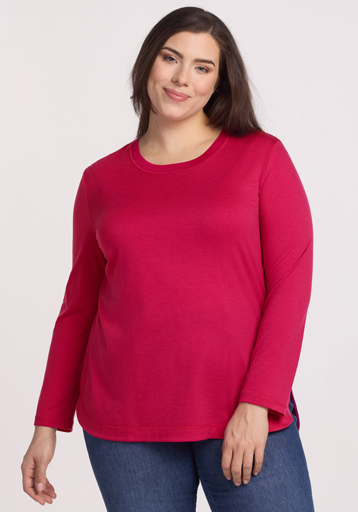 A woman with long dark hair is smiling softly, wearing the Woolx Hazel Tunic in Poppy Spark and blue jeans, as she stands against a plain light background. 