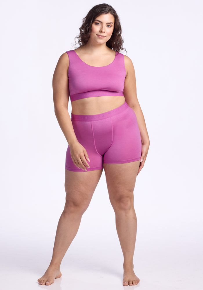 A woman with long, curly brown hair models the Woolx Elliott Boyshort - Wild Blossom, paired with a matching pink sports bra. She stands barefoot against a plain white background, smiling softly with one hand by her side.