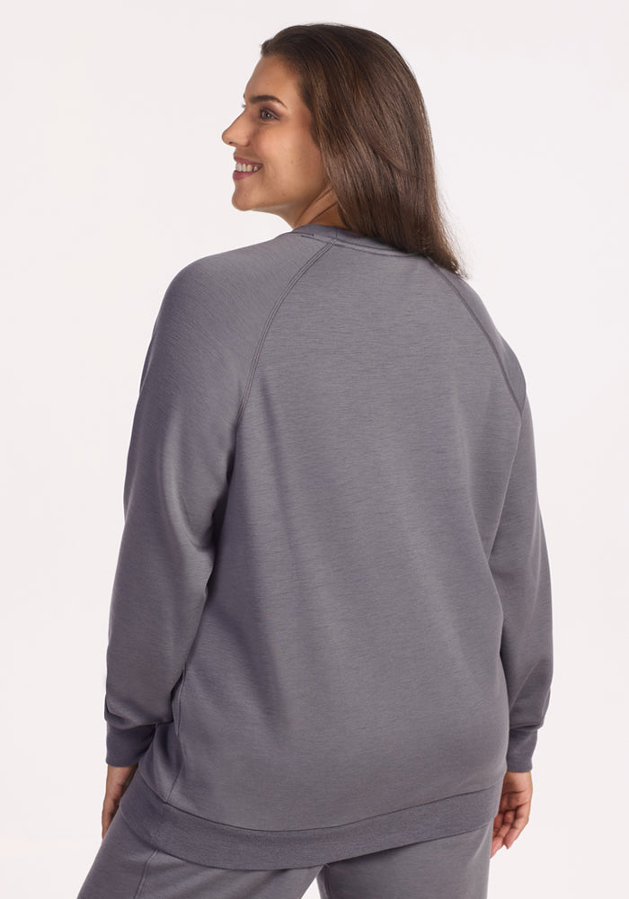 A woman with long brown hair smiles over her shoulder, wearing the Woolx Bailey Crewneck Sweatshirt in Arctic Dusk with matching pants, set against a plain, light-colored background.
