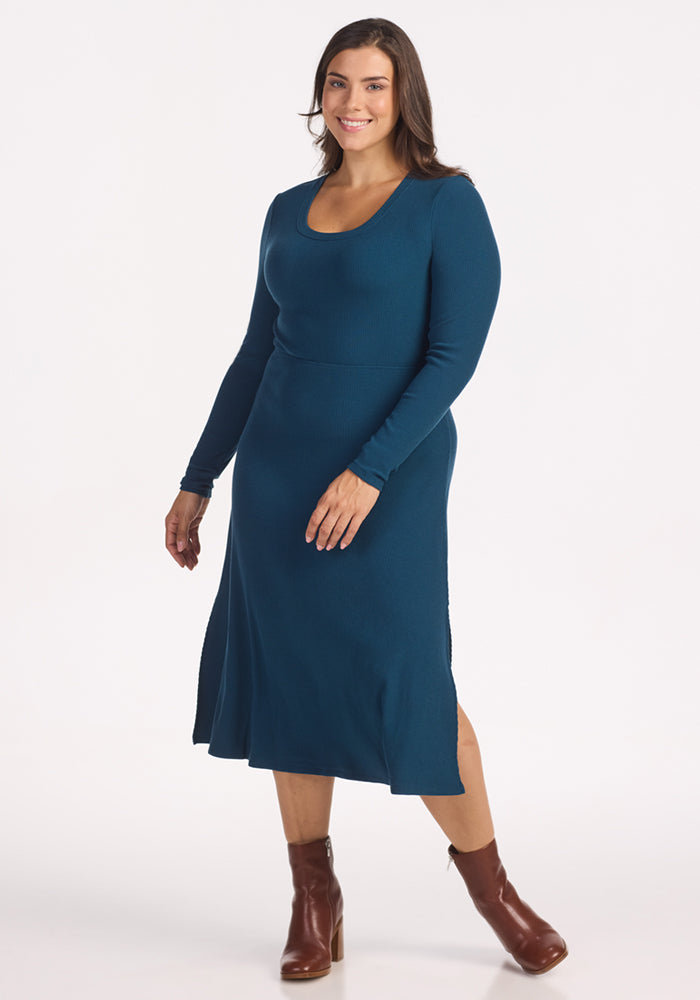 A woman with long brown hair smiles in the Woolx Caroline Ribbed Twirl Dress in Majolica Blue, featuring a scoop neckline and long sleeves. She pairs it with brown ankle boots and poses against a plain white background. 