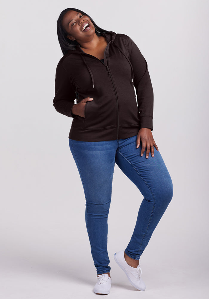 A woman wearing the Woolx Ryann Hoodie in French Roast, paired with blue jeans and white shoes, stands smiling with her head tilted back and one leg bent, posing cheerfully against a plain light background.