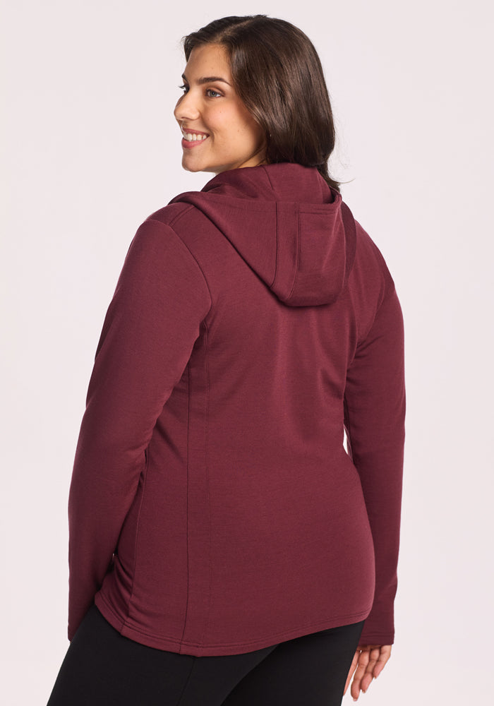A woman with medium-length brown hair smiles over her shoulder, wearing black pants and a Woolx Zoey Zip Up Hoodie in Toasted Garnet, standing against a plain light background.