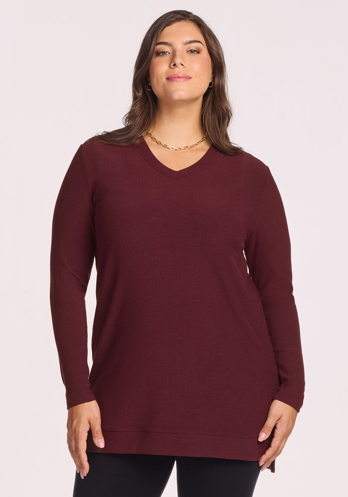 A woman with long brown hair wears the Woolx Evie Ribbed Tunic in Toasted Garnet and black pants, standing against a plain light background and looking confidently at the camera; this tunic offers style and temperature regulation. 