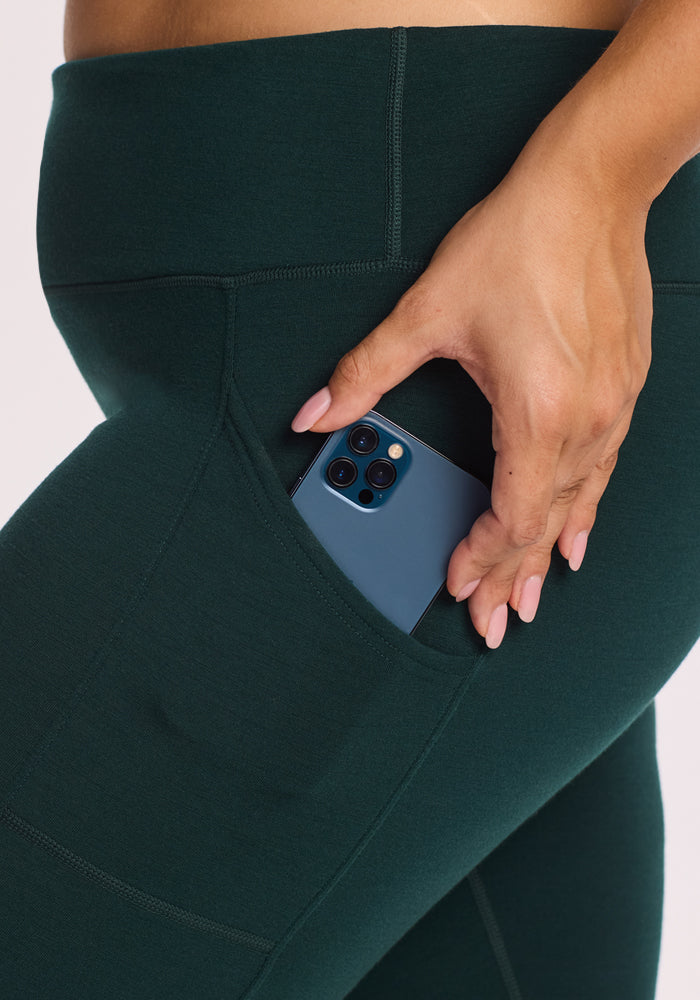 A woman wearing Woolx Piper Pocket Leggings in Deep Timber slips a blue smartphone into the side pocket, showcasing the baselayer’s convenient design and moisture-wicking Merino wool fabric.