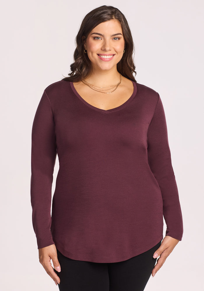 A woman with long brown hair smiles, wearing the Woolx Eva Tunic in Chocolate Fig and black pants, standing against a plain light background. 