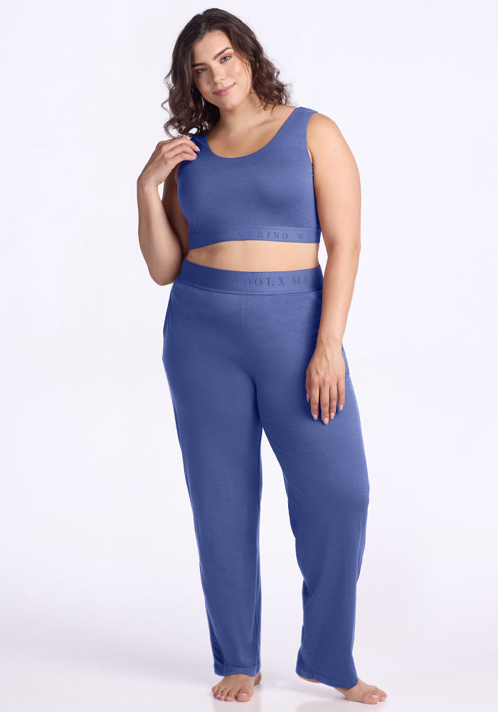 A woman with long brown hair wears the Woolx Aubrey Lounge Bra in Cloudy Bay, standing barefoot against a plain white background and smiling gently.