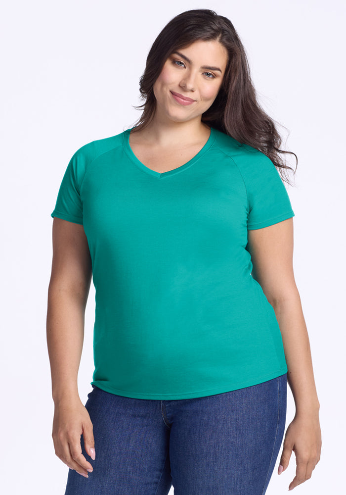A woman with long dark hair, wearing the Woolx Mia V Neck in Cool Jade and blue jeans, stands against a plain light background, smiling gently with one arm relaxed at her side. 