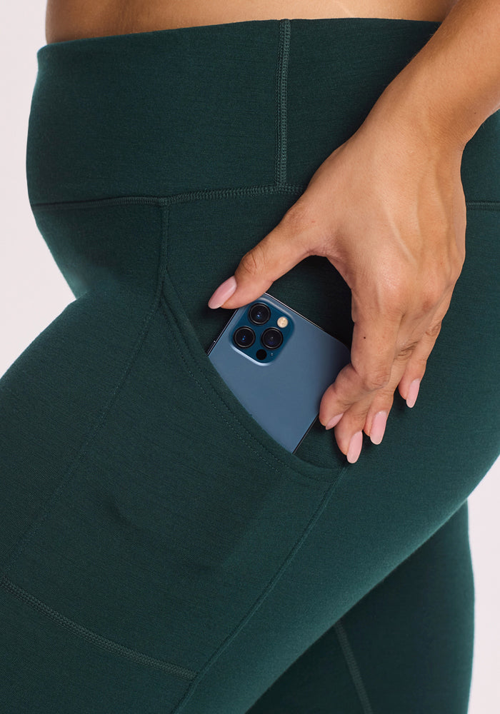 A close-up shows someone wearing Woolx Piper Pocket Leggings in Deep Timber, slipping a blue smartphone into the side pocket. Their hand is visible against a plain, light background.