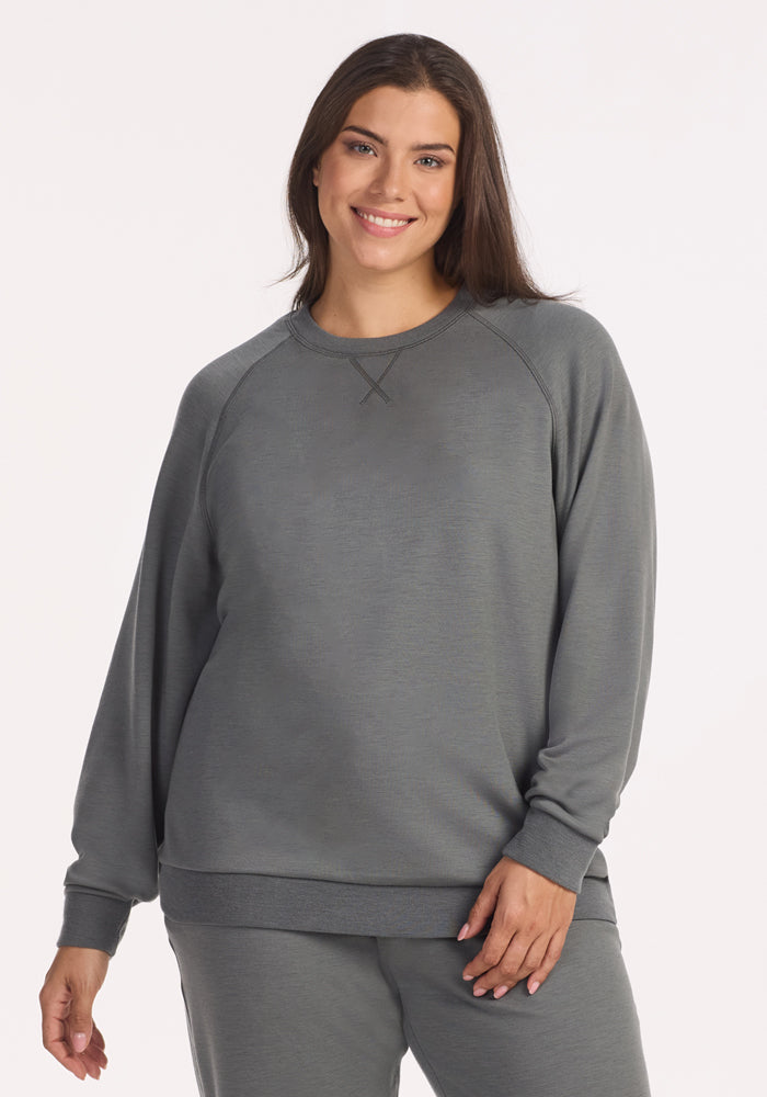 A woman with long brown hair smiles, wearing the Woolx Bailey Crewneck Sweatshirt in Sedona Sage and matching loose-fit pants, standing against a plain white background with one hand relaxed by her side. 