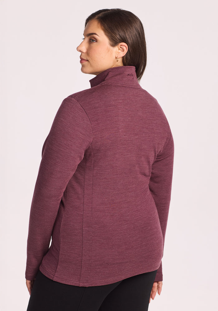 A woman with brown hair, wearing the Woolx Finley Zip Up Sweatshirt in Grape Harvest Melange and black pants, stands facing slightly away from the camera against a light background.