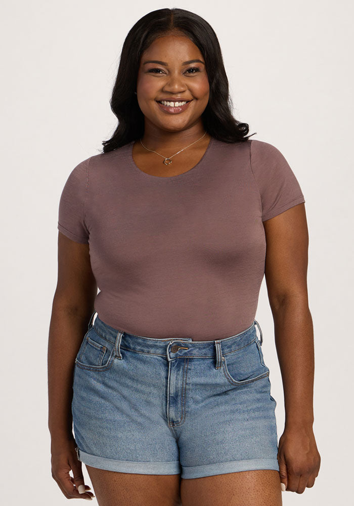A smiling woman in the Woolx Gracie Bodysuit - Peppercorn stands against a plain, light background. 