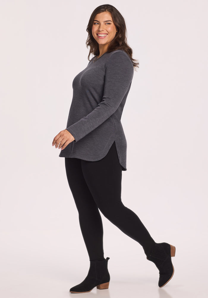 A dark-haired woman smiles in the Woolx Blair Tunic - Cinder Melange, paired with black leggings and ankle boots, enjoying lightweight warmth and odor-resistant comfort against a plain light background.