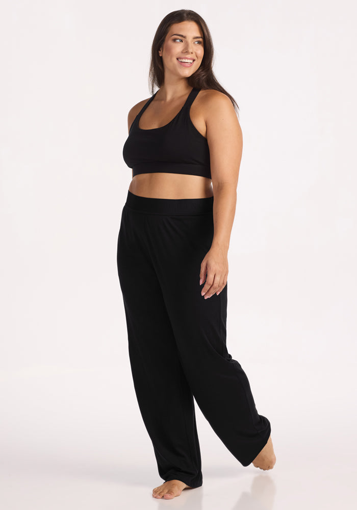 A woman stands barefoot, smiling confidently in Woolx Maya Lounge Pants - Black and a sports bra, posing against a plain white background.