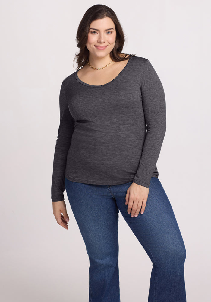 A woman with long brown hair smiles while wearing the Woolx Layla V Neck Top in Cinder Melange and blue jeans, standing against a plain light background.