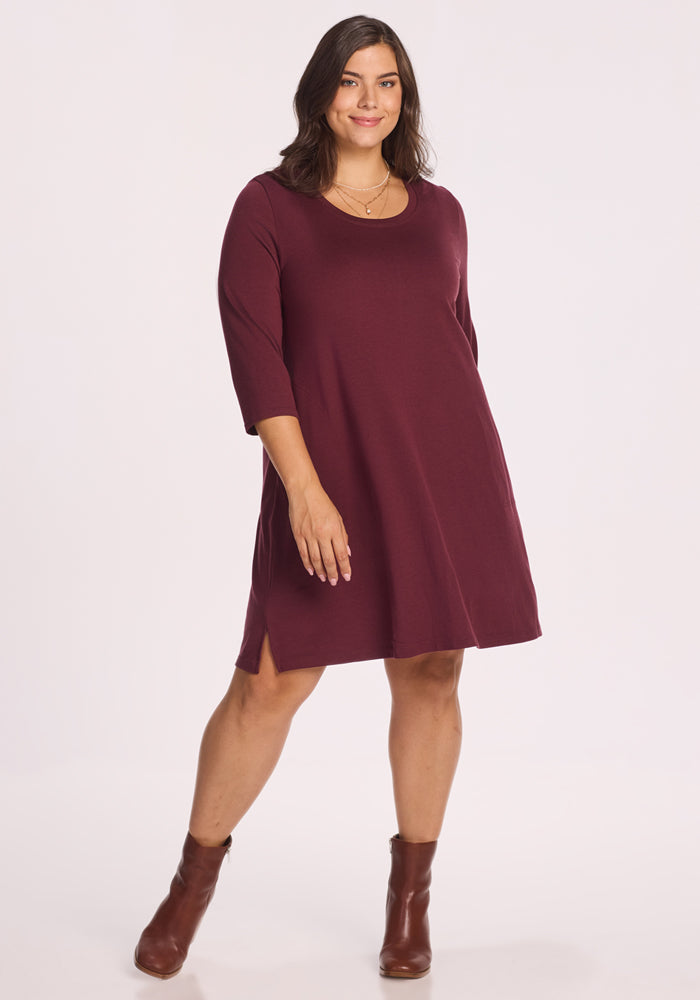 A woman smiles against a white background wearing the Woolx Rissa A Line Dress in Toasted Garnet with brown ankle boots—a knee-length, long-sleeve Merino wool style perfect for easy fall and winter layering. 