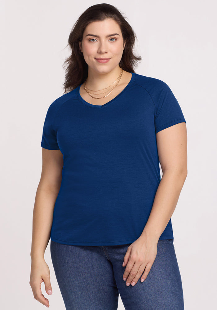 A woman with wavy brown hair wears the Woolx Mia V Neck in Estate Blue and blue jeans. She stands and smiles softly against a plain, light background. 