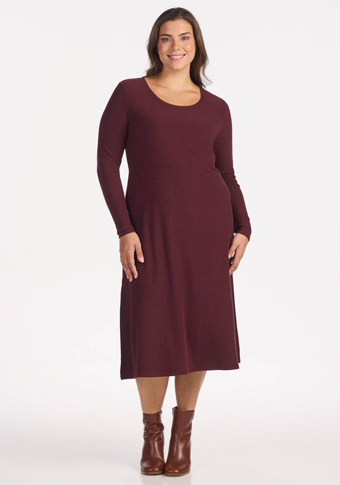 A woman with long brown hair models the Woolx Caroline Ribbed Twirl Dress in Toasted Garnet, featuring a wrinkle-resistant, versatile silhouette and paired with brown ankle boots, smiling against a plain white background. 
