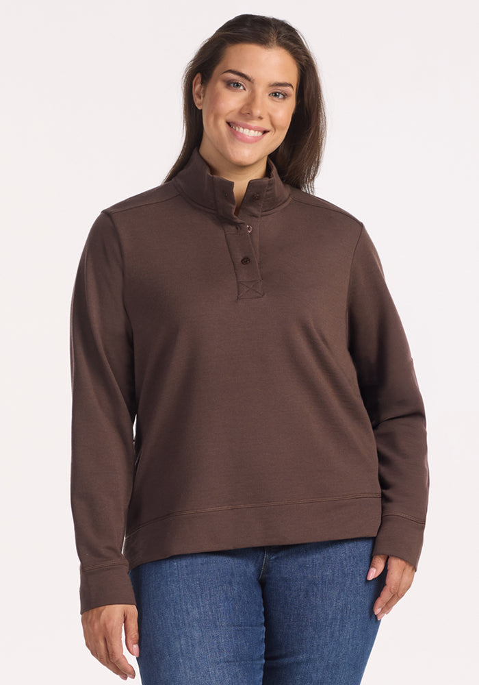 A woman with long brown hair smiles in the Woolx Annie 3 Button Pullover - Bracken and blue jeans, standing against a plain light background. 