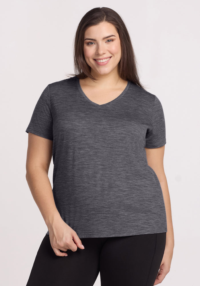 A woman with long brown hair smiles, wearing a Woolx Junie V Neck in Cinder Melange and black pants. Against a plain light background, she stays comfortable all day thanks to the tee’s moisture-wicking Merino wool fabric. 