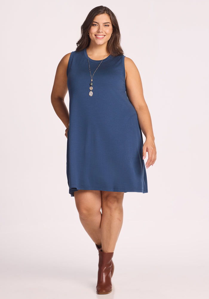A woman with long hair and a long necklace smiles confidently in the Woolx Clara Dress - Starry Night, a sleeveless, knee-length blue merino wool dress, paired with brown ankle boots against a plain, light background. 