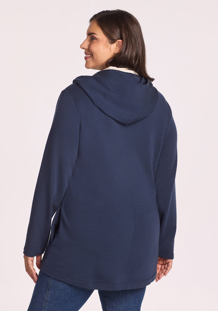 A woman with shoulder-length brown hair, wearing the Woolx Edna Hooded Cardigan in Deep Navy and blue jeans, stands smiling and faces away from the camera against a light background.